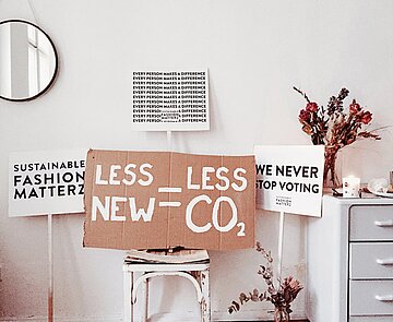 Demoplakate in einem Zimmer, auf einer zentral positionierten Pappe steht: Less New - Less CO2